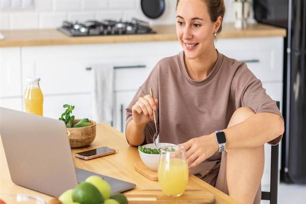 Ayuno intermitente - Mujer comiendo saludable en la cocina de su casa viendo algo en la pantalla de su portátil