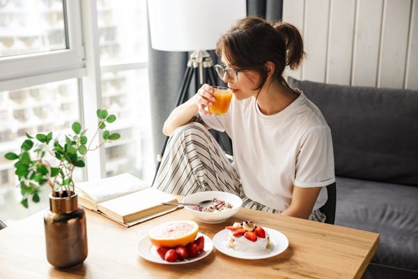 Mujer tomando desayuno saludable en la mesa del salón mientras lee un libro