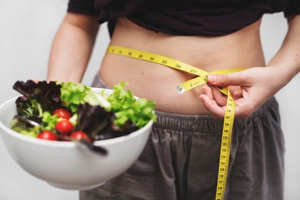 Fascitis plantar - una mujer con una ensalada en la mano derecha mientras se mide la cintura con un cinta métrica con la mano izquierda