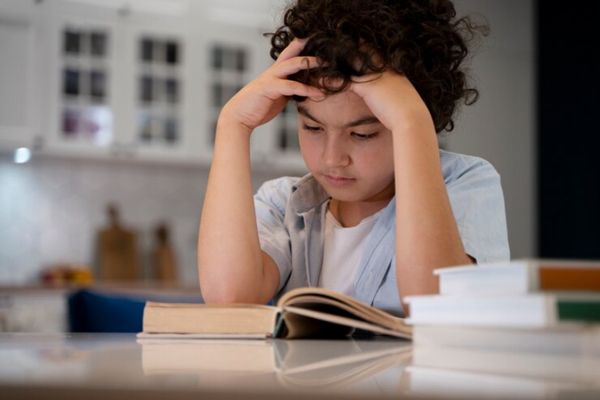 Afasia - niño leyendo un libro sobre la mesa, llevándose las manos a la cabeza porque no entiende bien lo que lee