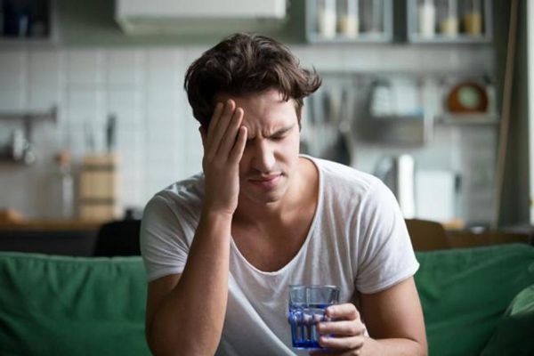 Ayuno intermitente - Hombre con dolor de cabeza sentado en el sofá con un vaso de agua en su mano
