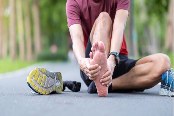 Fascitis plantar - hombre sentado en el camino de un parque agarrándose con fuerza la planta de su pie derecho desnudo