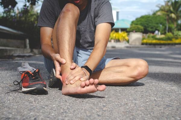 Esguince de tobillo - hombre sentado en el camino de un parque agarrándose el pie desnudo a la altura del tobillo por un posible esguince de tobillo