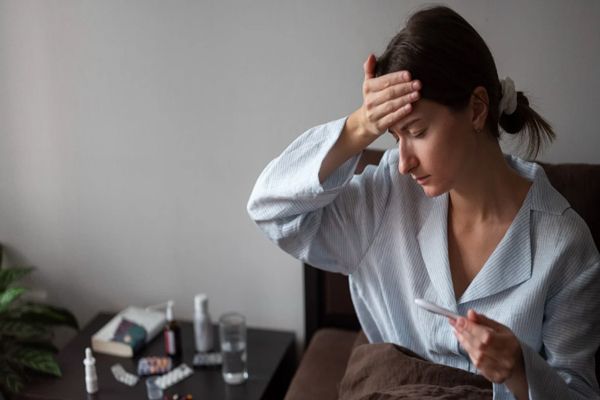 COVID19 - mujer blanca en pijama dentro de la cama tomándose la temperatura con una gran cantidad de medicamentos en la mesilla de noche