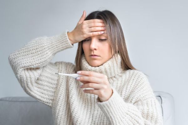 Lupus - mujer blanca con el pelo largo y con un jersey de punto de cuello largo sentada en un sofá mientras mira un termómetro