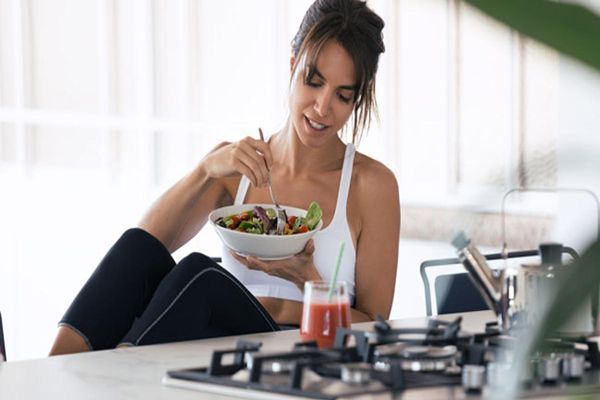 Ayuno intermitente - Mujer vestida con ropa deportiva sentada al lado de la cocina comiendo una ensalada y un zumo