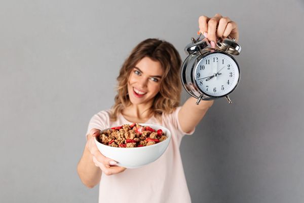 Ayuno intermitente - Mujer contenta con un bol de cereales y fresas en la mano derecha y un despertador en la mano izquierda