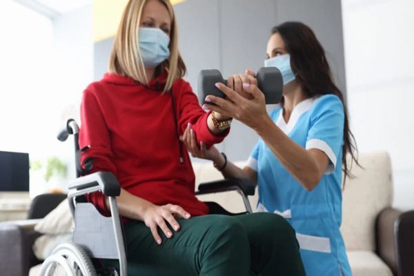 mujer en sillas de ruedas haciendo rehabilitación para ganar fuerza en los brazos con la ayuda de una enfermera en su casa