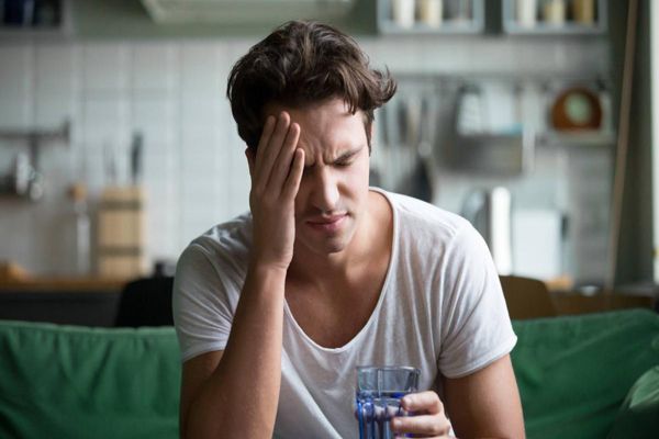 Tuberculosis - hombre sentado en el sofá con fuertes dolores de cabeza con un vaso de agua en la mano