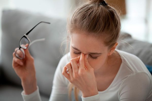 Esclerosis múltiple - mujer con las gafas en la mano y mientras la otra mano acaricia con los dedos la zona del puente de la nariz con los ojos en señal de cansancio