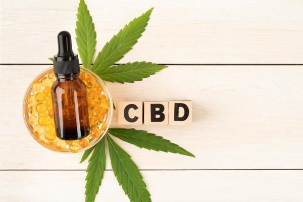 producto con cannabidiol en formato de aceite dentro de un frasco con cuentagotas y en capsulas, junto a unas hojas cáñamo y las siglas del principio activo