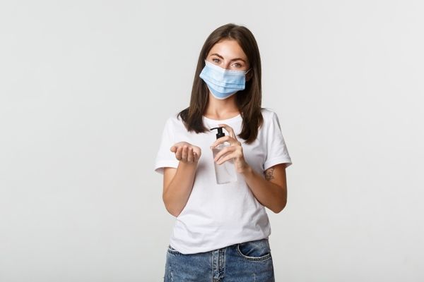 mujer blanca con vaqueros y camiseta blanca, usando mascarilla y lavando sus manos con gel hidro alcohólico