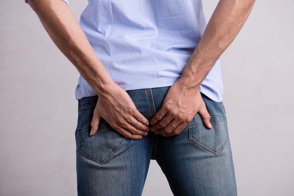Hemorroides - hombre con vaqueros y una camiseta azul agarrándose el trasero por los dolores que le causan la hemorroides