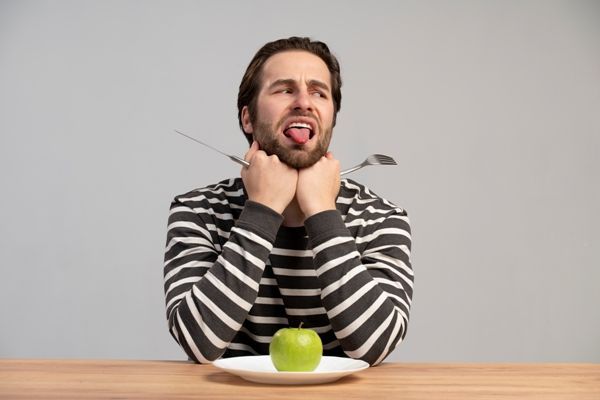 Disfagia - hombre adulto sentado en la mesa con un plato donde hay una manzana, con las manos cerradas sujetando un cuchillo en la derecha y el tenedor en la izquierda. Apoyando la cabeza en los puños porque no puede tragar