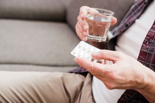 hombre sentado en un sofá con un blíster de pastillas en la mano izquierda y un vaso de agua en la mano derecha