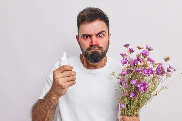 Alergias - Hombre con rinitis producida por su alergia al polen de las flores que tiene en la mano izquierda mientras sujeta en su mano derecha un medicamento para la alergia por vía nasal