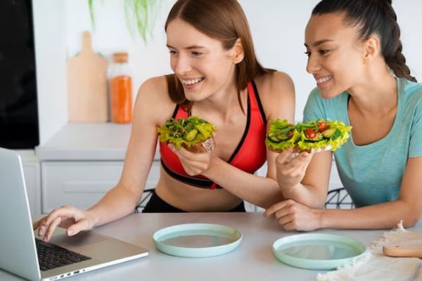 Amigas en la cocina viendo algo en la pantalla del portátil mientras comen algo saludable