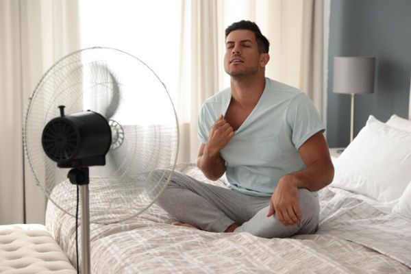 Hiperhidrosis - Hombre en pijama sentado sobre la cama enfrente de un ventilador de pie para refrescarse