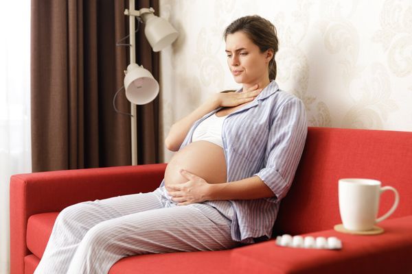 Hipertiroidismo - mujer embarazada sentada en el sofá con una taza y medicamentos para tratar problemas de tiroides llevándose la mano a la garganta