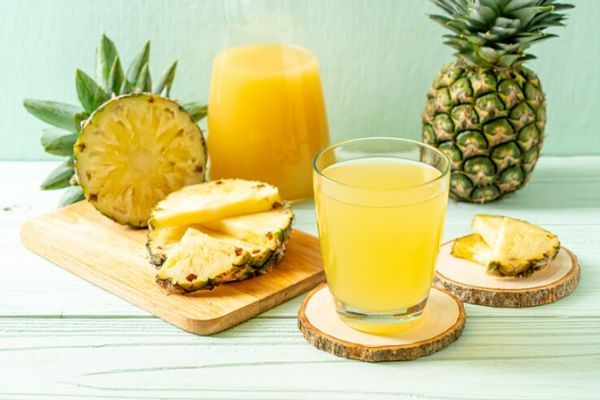 Mesa con una piña entera y otra cortada en gajos, además de una botella y un vaso llenos del zumo de esta fruta rica en Bromelia