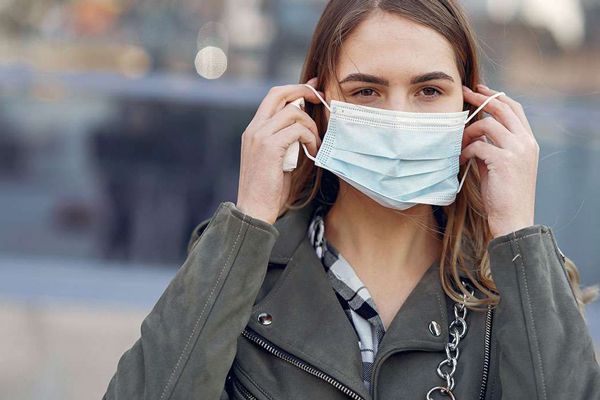 Mujer con camisa de cuadros y camisa de ante caminando por la calle mientras se pone una mascarilla contra los alérgenos en el aire