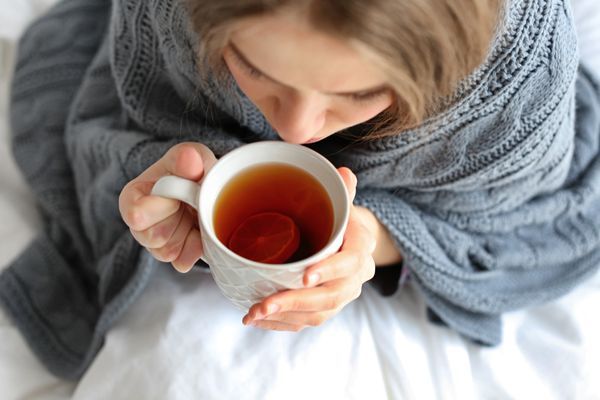 mujer en la cama con una manta sobre los hombres tomando una taza de té con una rodaja de limón