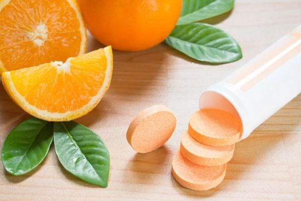 Mesa con una naranja entera, otra cortada por la mitad y un gajo de la fruta al lado de un bote de pastillas con vitamina C