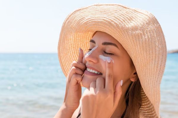 mujer con sombrero en la playa poniéndose crema en la cara