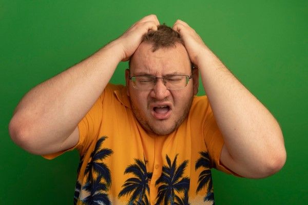 Hombre molesto con gafas y camisa naranja que parece molesto e irritado con las manos en la cabeza de pie sobre la pared verde