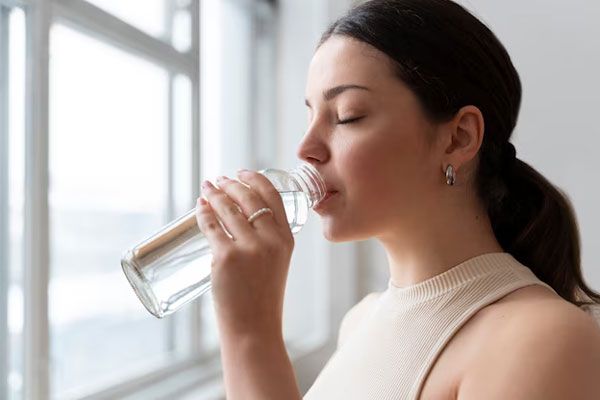 Mujer con piel saludable bebé un abundante vaso de agua junto a la ventana de su casa 