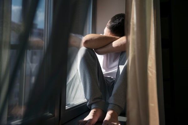 Trastorno de ansiedad - hombre sentado en la ventana con las rodillas en el pecho y los brazos cruzados delante de la cara