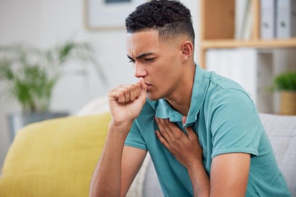 bronquitis crónica - joven sentado en el sofá tosiendo mientras se lleva la mano al pecho