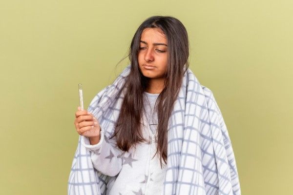 Fiebres de origen desconocido - joven en pijama con una manta sobre los hombros y mirando un termómetro en la mano derecha por si tiene fiebre