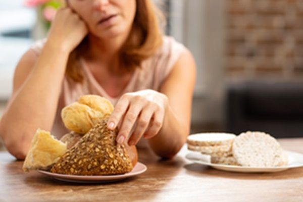 Celiaquía o intolerancia al gluten - Mujer agarrando una pieza de pan mientras tiene varios alimentos con gluten sobre la mesa