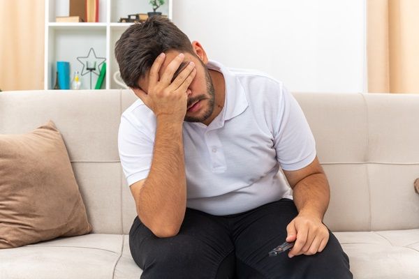 Hombre joven sentado en un sofá poniéndose la mano en la cara con gestos de tristeza y depresión