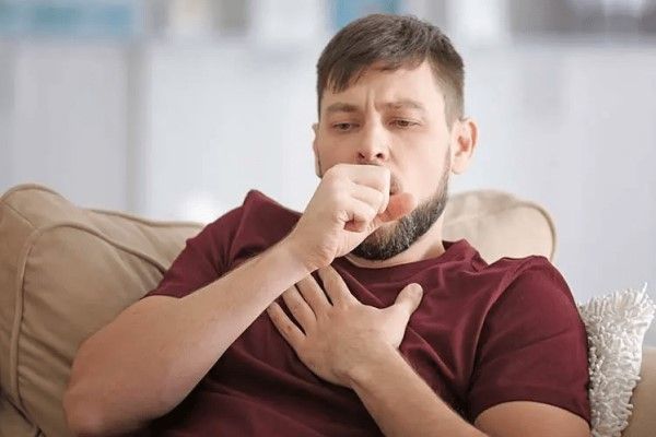 Bronquitis sin fiebre - hombre en el sofá tosiendo mientras se agarra el pecho