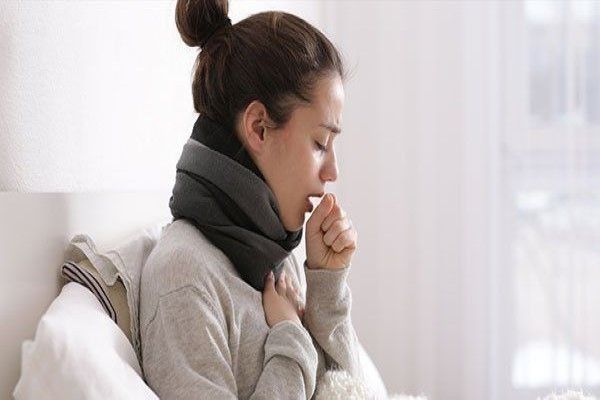 Bronquitis sin fiebre - mujer en la cama con una bufanda tosiendo mientras se agarra el pecho