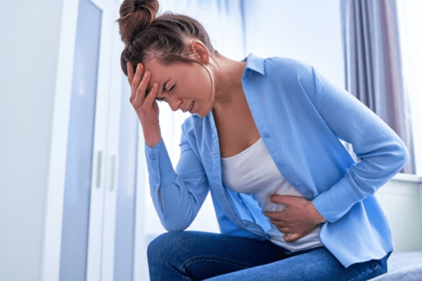Mujer joven sentada, con una mano en el abdomen y la otra en la cabeza, mostrando posibles molestias digestivas
