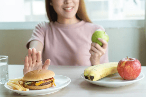 Colesterol bueno y malo - Mujer joven rechazando una hamburguesa y eligiendo una fruta como opción saludable para equilibrar el colesterol bueno y malo