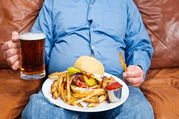 Colesterol bueno y malo - Hombre con sobrepeso sosteniendo comida chatarra y cerveza, destacando cómo las grasas saturadas afectan el colesterol