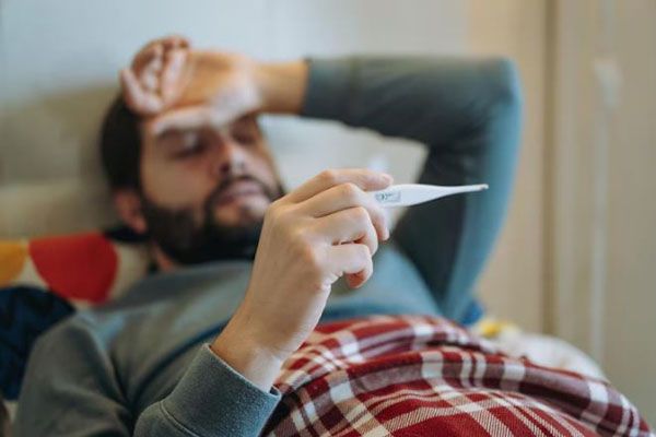 Cómo bajar la fiebre - Hombre acostado se toma la temperatura corporal con un termómetro digital