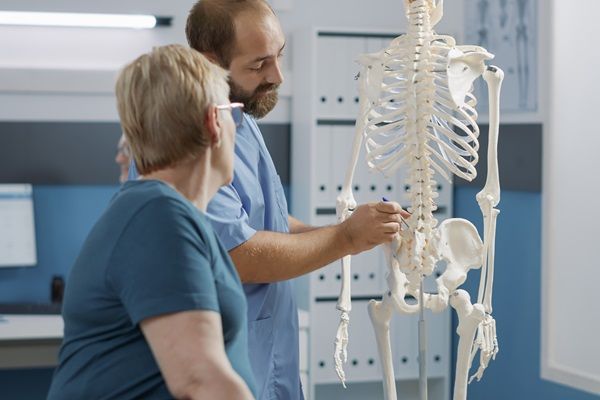 Qué es la osteoporosis - Doctor señalando en un esqueleto dónde se encuentra la afección de la paciente, la cual sufre de osteoporosis