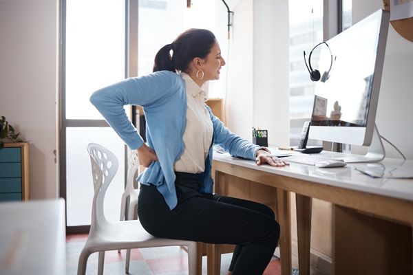 Joroba - Mujer con fuerte dolor de espalda al estar sentada trabajando, debido a la afección que presenta en la columna vertebral por la mala postura